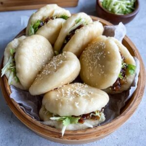 Bao buns with pork filling on a bamboo steamer, white pillowy soft buns split open with glossy hoisin-glazed pork belly slices, pickled cucumber, fresh cilantro, sliced scallions, sesame seeds